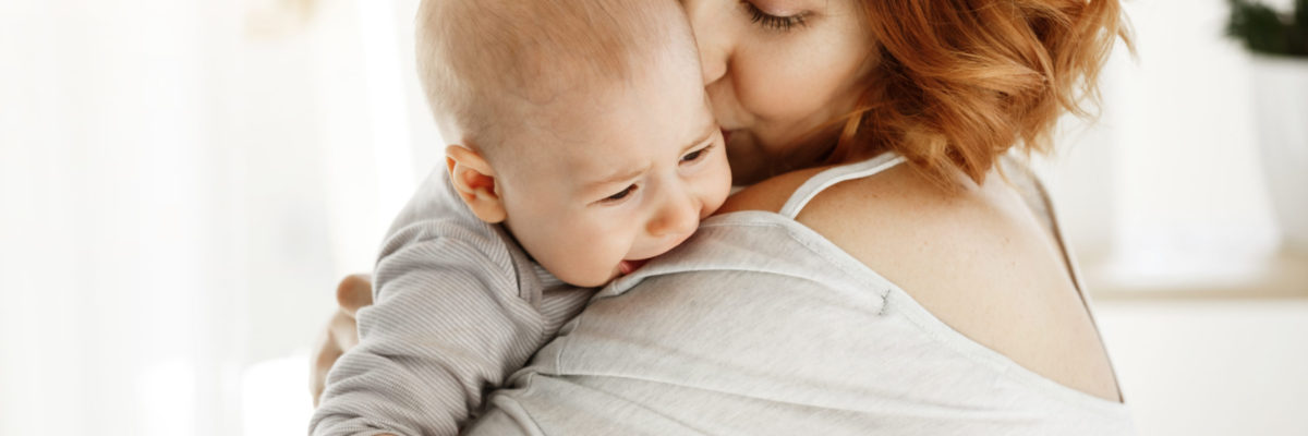 Young good looking mother hugs and calms her crying baby. Child screaming and crying on mother shoulder. Scene of protection and love. Family concept